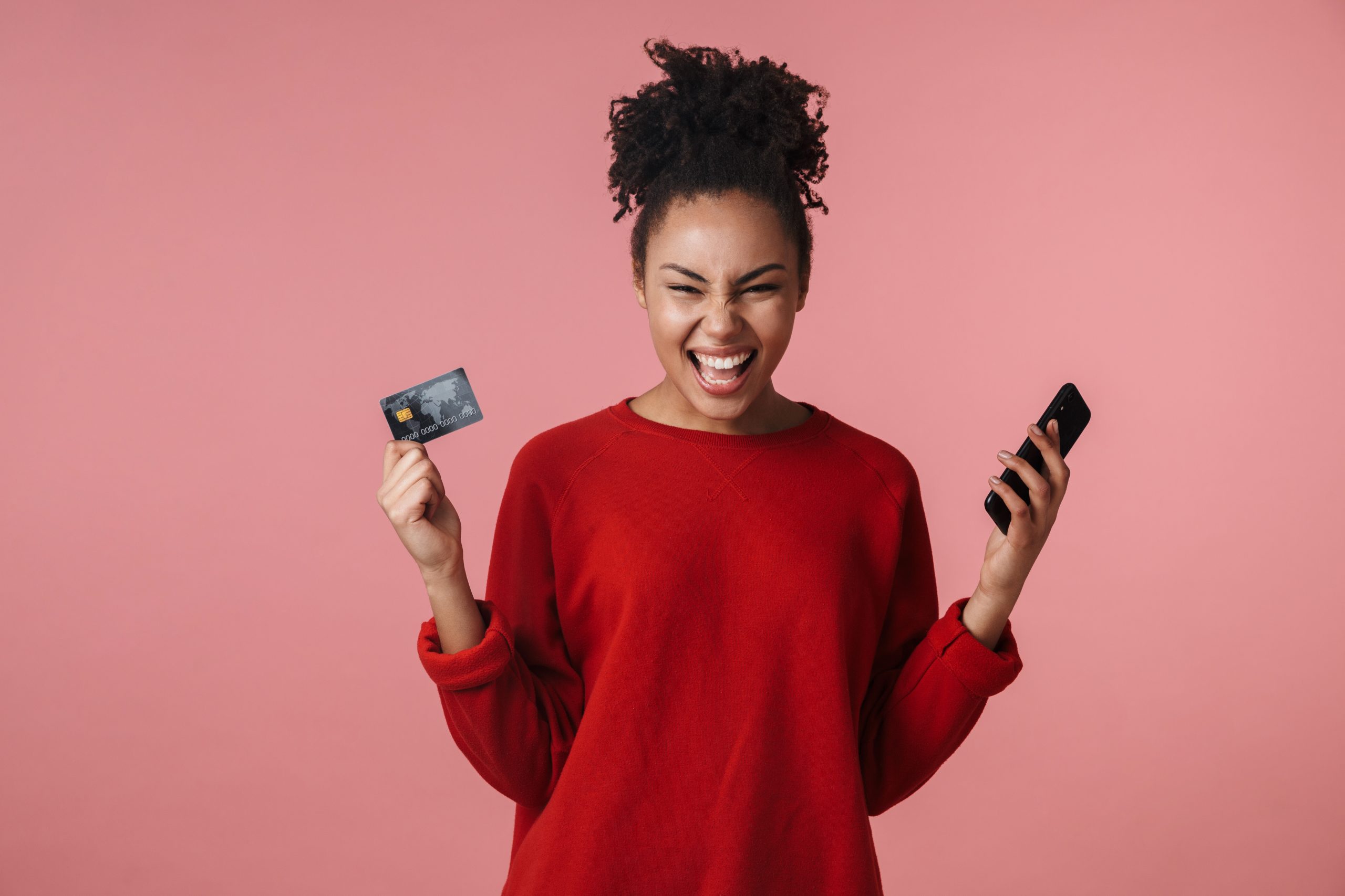 Amazing shocked excited young african woman posing isolated over pink wall background using mobile phone make winner gesture holding credit card.