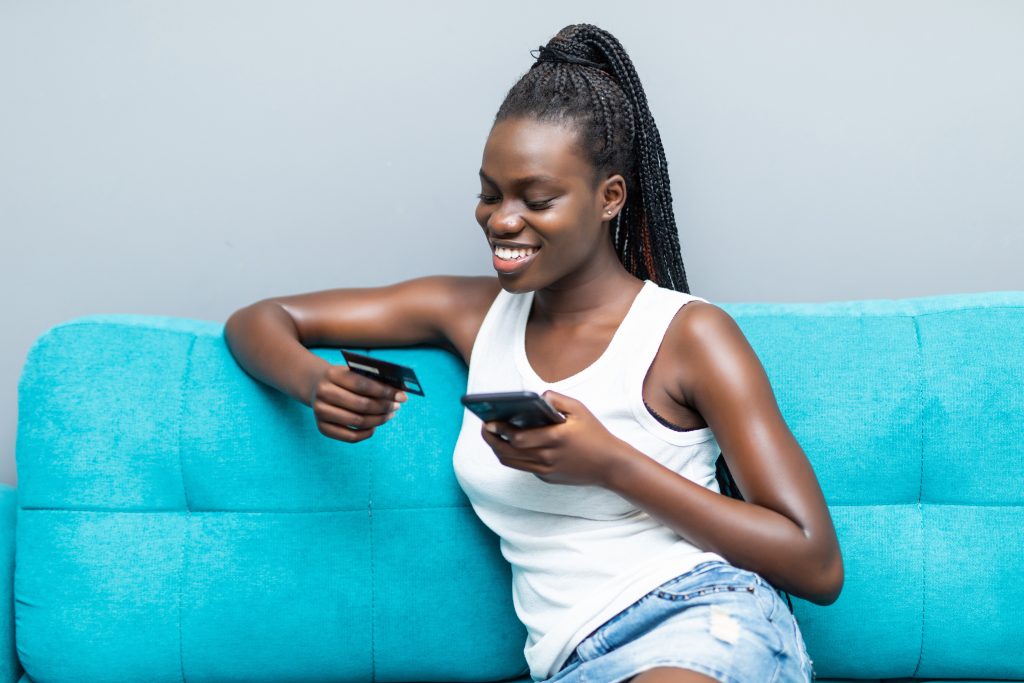 African American woman using credit card for buying and banking online with mobile phone sitting on couch at home