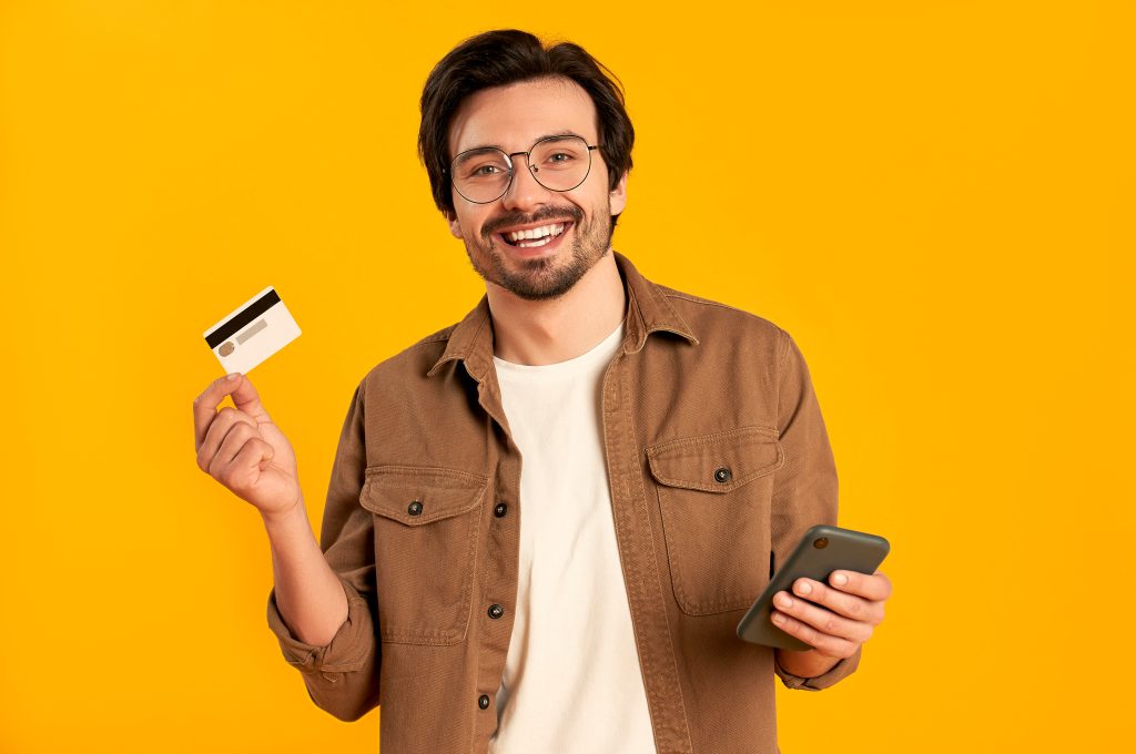 Young bearded man in glasses holds credit card and smartphone is