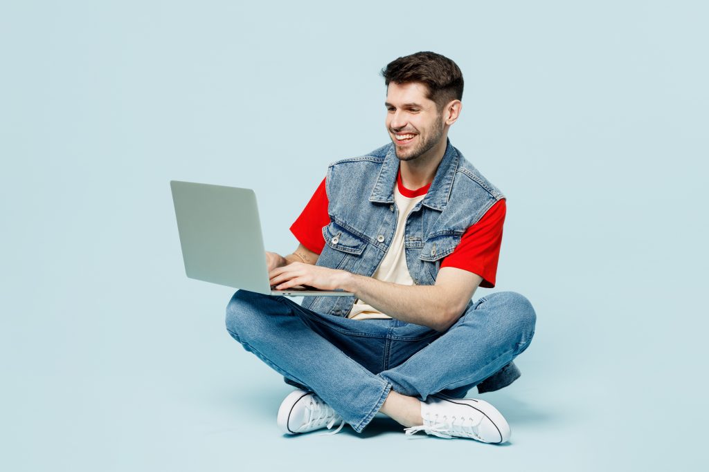 Full body smiling fun young happy IT man wears denim vest red t-shirt casual clothes sit hold use work on laptop pc computer chatting online isolated on plain pastel light blue cyan background studio.