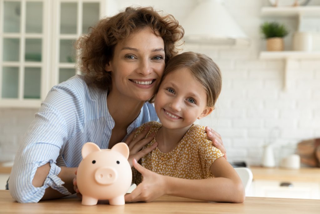 Portrait of affectionate mother and daughter sitting at table with piggybank.